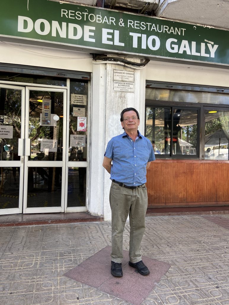 Letrero: Restobar y restaurant DONDE TÍO GALLY. Fotografía completa de persona que está de pie con camisa, pantalón y zapatos. Su nombre es tío gally, dueño del local de comida típico de la comuna de Peñaflor, Santiago de chile.